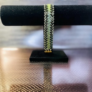 Op de afbeelding: Een zwarte en groene kralen armband met een geometrisch patroon. De armband wordt getoond op een zwarte standaard.
