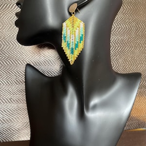May include: Geometric beaded earrings in yellow, teal, and white. The earrings are a long, hexagonal shape with a gold-coloured post. The earrings are displayed on a black mannequin head.