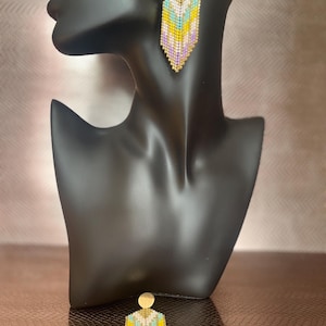 May include: Geometric beaded earrings in shades of yellow, blue, white, and lavender. The earrings have a gold-coloured top and bottom, and are displayed on a black mannequin head. The earrings are approximately 7.6 cm long.