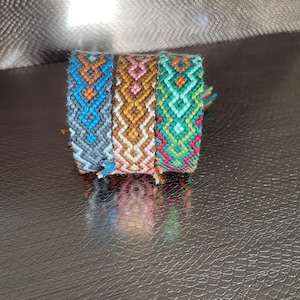 Könnte beinhalten: Vier Freundschaftsbänder, die übereinander gestapelt sind. Die Armbänder sind aus buntem Stickgarn gefertigt und haben geometrische Muster. Die Armbänder sind blau, braun, rosa und grün.