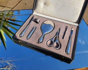 May include: A silver-coloured vanity set in a black leatherette case. The set includes a mirror, scissors, tweezers, a nail file, a cuticle pusher, and a thimble.