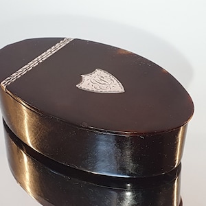 以下が含まれることがあります： 蓋に銀の盾が付いた楕円形の木製箱。盾にはモノグラムが刻まれています。