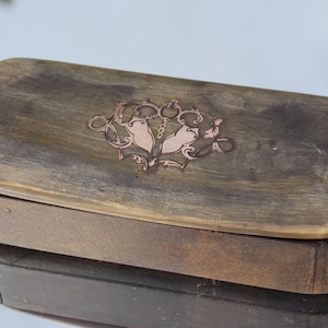 Puede incluir: Una caja de madera con tapa abatible. La tapa está decorada con un diseño tallado de dos pájaros estilizados con una corona encima. La caja es marrón y tiene un aspecto desgastado y antiguo.