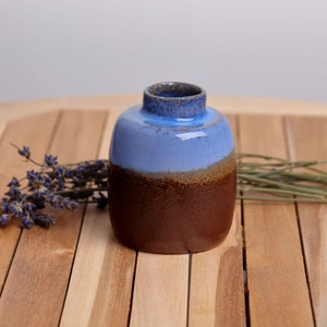 Pode incluir: Um pequeno vaso de cerâmica artesanal com um corpo arredondado e um gargalo estreito. O vaso é esmaltado em um esquema de duas cores, com uma metade superior azul claro e uma metade inferior marrom escuro. Exibido em uma superfície de madeira.