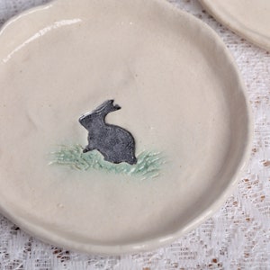 Può includere: Un piatto in ceramica bianco sporco con un coniglio grigio su erba verde. Il piatto ha una forma irregolare e una finitura liscia. Perfetto per servire antipasti o piccoli piatti.
