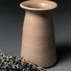 Op de afbeelding: Een beige keramische vaas met een getextureerd oppervlak en een uitlopende rand. De vaas wordt getoond met een takje groen met kleine blauwe en witte bloemen. De vaas heeft een neutraal kleurenpalet.
