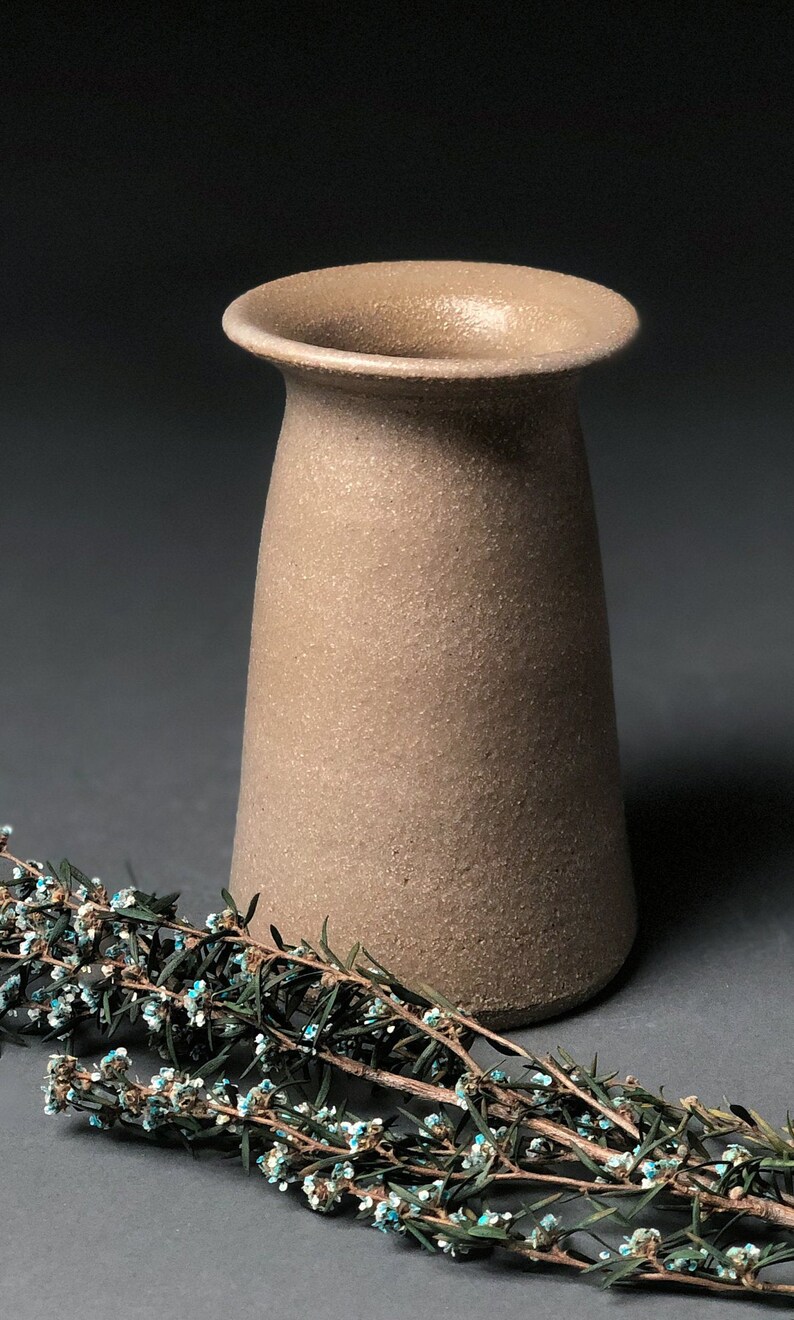 Op de afbeelding: Een beige keramische vaas met een getextureerd oppervlak en een uitlopende rand. De vaas wordt getoond met een takje groen met kleine blauwe en witte bloemen. De vaas heeft een neutraal kleurenpalet.
