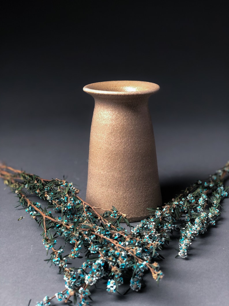 Op de afbeelding: Een beige keramische vaas met een getextureerd oppervlak. De vaas heeft een uitlopende rand en een licht taps toelopend lichaam. Takken met kleine blauwe en witte bloemen zijn voor de vaas geplaatst.