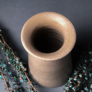 Op de afbeelding: Een beige keramische vaas met een brede, uitlopende opening en een getextureerd oppervlak. De vaas heeft een cilindrisch lichaam en staat op een donkere achtergrond, met takjes blauwe en groene bloemen eromheen.