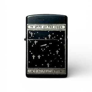 May include: Black lighter with a starry night design. The lighter features the text "IN SPITE OF THE ODDS" at the top and "I'M GLAD YOU FOUND ME" at the bottom. The design includes stars and crosses.