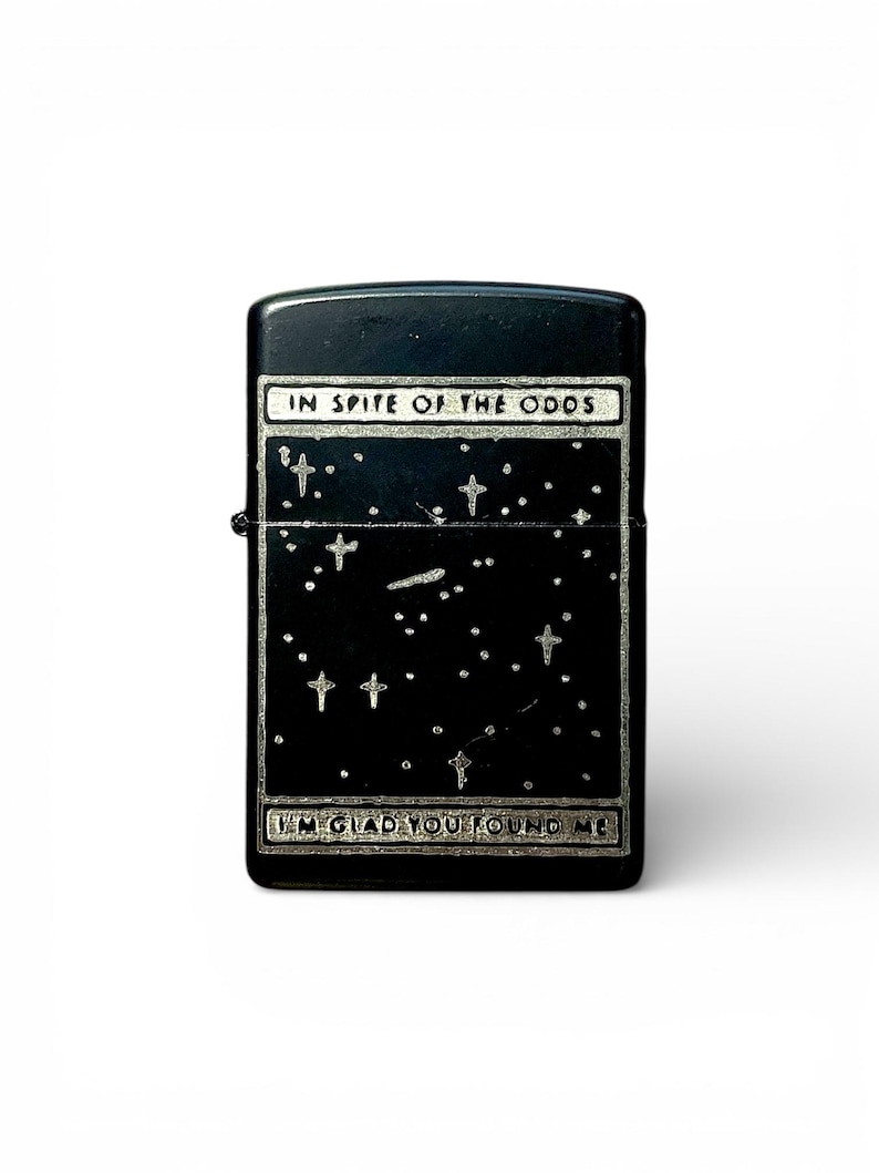 May include: Black lighter with a starry night design. The lighter features the text "IN SPITE OF THE ODDS" at the top and "I'M GLAD YOU FOUND ME" at the bottom. The design includes stars and crosses.