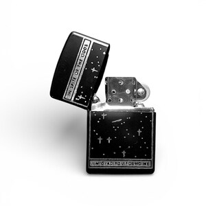 May include: Black and silver lighter with the lid open, displaying the text "IN SPITE OF THE ODDS" and "I'M GLAD YOU FOUND ME". The lighter has a starry night design with small crosses.