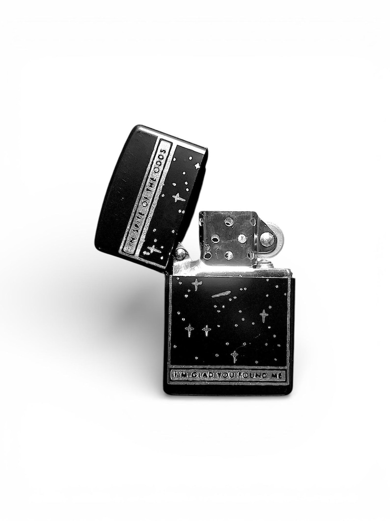 May include: Black and silver lighter with the lid open, displaying the text "IN SPITE OF THE ODDS" and "I'M GLAD YOU FOUND ME". The lighter has a starry night design with small crosses.