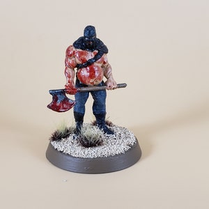 May include: A miniature figurine of a muscular figure with a black hood and blood-stained torso, holding a blood-covered axe. The figure stands on a circular base with small plants and gravel.