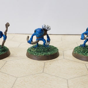 May include: Three blue frog figurines, each with a different expression and holding a different object. The frog on the left is holding a stick, the frog in the middle is holding a curved stick and a small white object, and the frog on the right is holding a small knife.