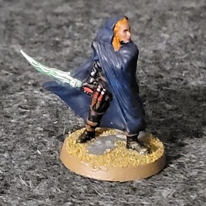 Puede incluir: Una figura en miniatura de una persona que lleva una capa azul y sostiene una espada verde brillante. La figura está de pie sobre una base marrón.