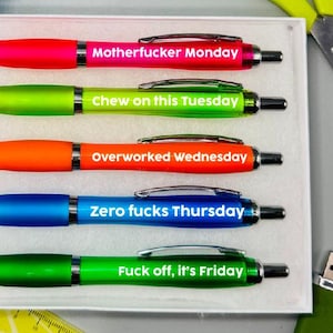 May include: A set of five colorful pens with humorous phrases for each weekday, including "Motherfucker Monday" and "Fuck off, it's Friday." The pens are in a white box, with a blue stapler, yellow scissors, and a green USB drive nearby.