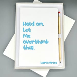 Peut inclure: Un carnet blanc avec le texte "Hold on. Let me overthink this." en lettres bleues. Le carnet est dans une boîte blanche avec un crayon.