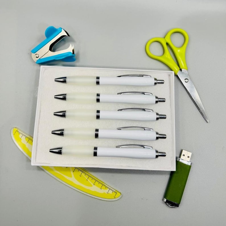 May include: An assortment of office supplies including five white pens in a box, a blue stapler, yellow scissors, a yellow ruler, and a green USB drive. The pens have silver accents and black ink tips. The scissors have yellow handles.