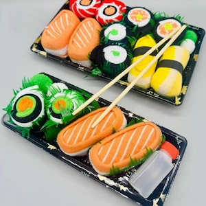 May include: Two trays of sushi, with various rolls and nigiri. The sushi includes orange, white, and green elements, with wooden chopsticks and a small bottle of sauce. The sushi is arranged on black trays with a gold pattern.