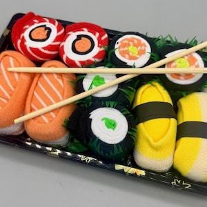 Pode incluir: Um conjunto decorativo de sushi disposto numa bandeja preta com pauzinhos de madeira. O sushi inclui peças laranja, vermelhas, pretas e amarelas, imitando vários tipos de sushi. O conjunto foi concebido para se assemelhar a uma refeição japonesa.