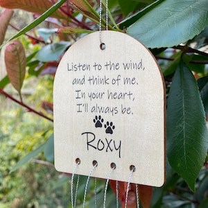 Pode incluir: Carrilhão de vento de madeira com uma placa em forma de coração que diz "Ouça o vento, e pense em mim. No seu coração, eu sempre estarei." e o nome "Roxy" abaixo. O carrilhão tem uma corrente de contas pendurada.
