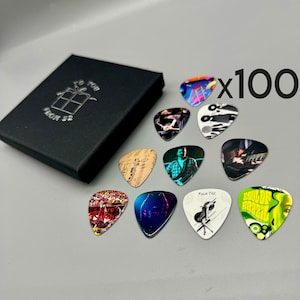Puede incluir: Un conjunto de 100 púas de guitarra con varios diseños, incluyendo notas musicales, una persona tocando la guitarra y un diseño abstracto colorido. Las púas se muestran en una caja negra con el texto "To You From Sz" en la parte superior.