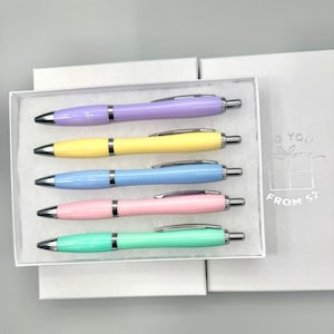 May include: A set of five pastel-colored pens in a white box. The pens are light purple, yellow, blue, pink, and green. Each pen has a silver clip and tip. The box is inside a larger white box with the text "TO YOU" and "FROM 52".