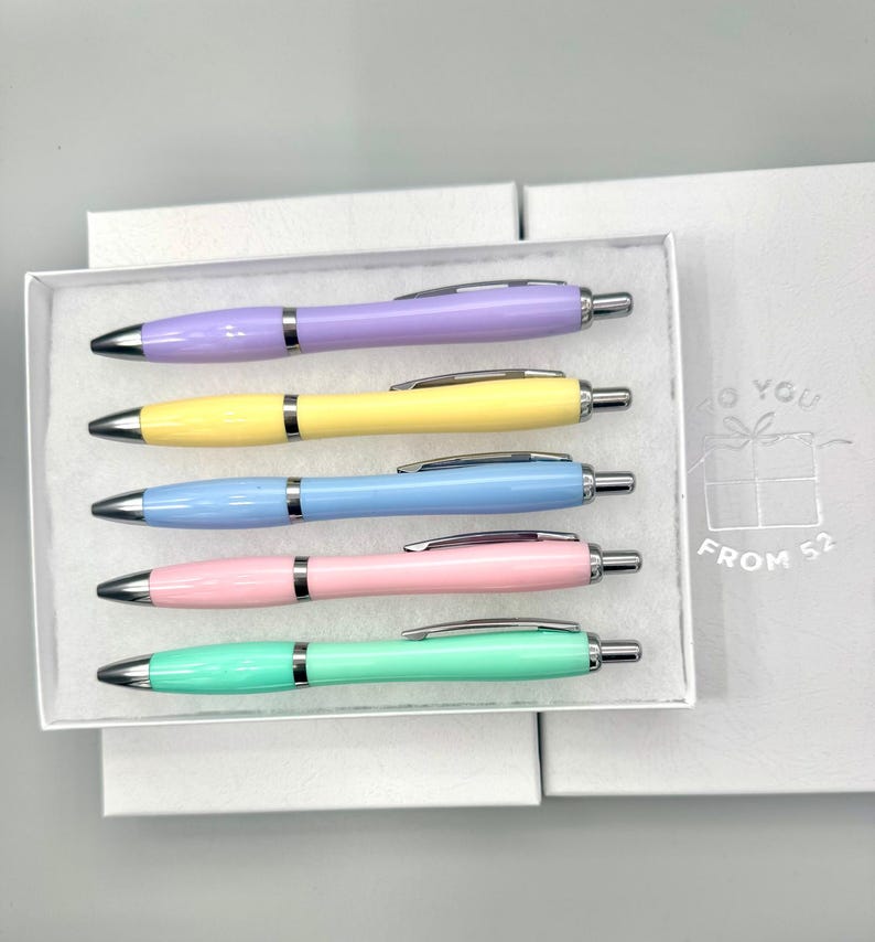May include: A set of five pastel-colored pens in a white box. The pens are light purple, yellow, blue, pink, and green. Each pen has a silver clip and tip. The box is inside a larger white box with the text "TO YOU" and "FROM 52".
