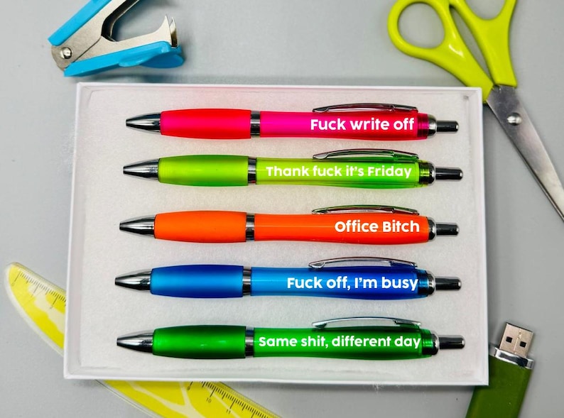 Op de afbeelding: Een set van vijf kleurrijke pennen met zwarte inkt, elk met een andere zin in witte tekst. De pennen liggen in een witte doos, met een nietmachine, een schaar, een liniaal en een USB-stick op de achtergrond.