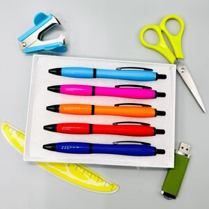 May include: A set of five colorful pens in a white box, with a blue stapler, yellow scissors, a green USB drive, and two yellow rulers. The pens are blue, pink, orange, red, and dark blue. The pens have black ink and black accents.