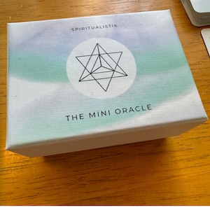 Könnte beinhalten: Eine weiße Box mit einem aquamarinen und violetten Aquarelldesign. Die Box hat einen schwarzen Davidstern mit einem kleineren Dreieck im Inneren. Der Text "SPIRITUALISTIK THE MINI ORACLE" ist auf der Box aufgedruckt.