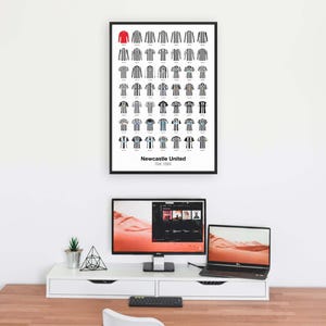 May include: A framed print displaying a series of Newcastle United football shirts, predominantly black and white striped. A single red shirt is at the top. The print is above a desk with a computer, laptop, and other office items.