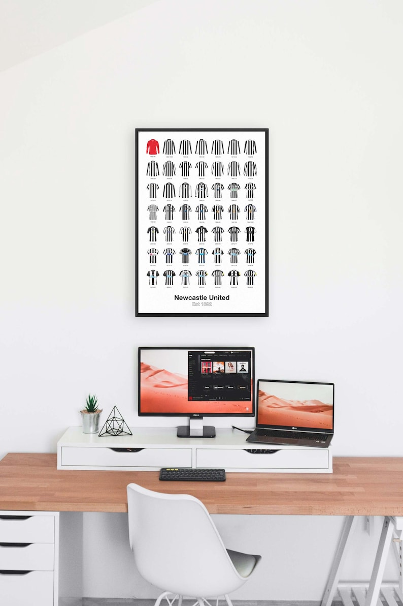May include: A framed print displaying a series of Newcastle United football shirts, predominantly black and white striped. A single red shirt is at the top. The print is above a desk with a computer, laptop, and other office items.