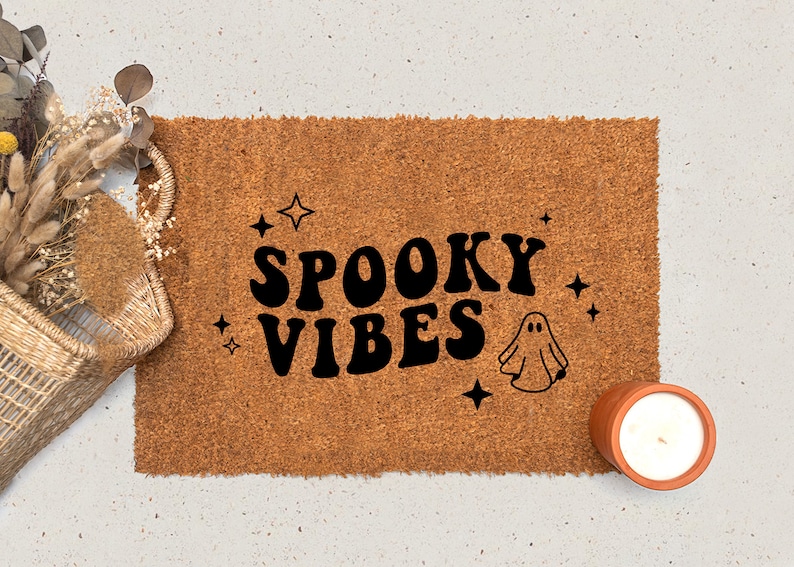 May include: A brown coir doormat with the text "SPOOKY VIBES" in black retro font. A small white ghost illustration is to the right of the text. There are small white stars scattered around the text and ghost.