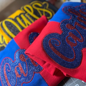 May include: Close-up of folded sweatshirts in blue, red, and black. Each sweatshirt features a glittery script design. The blue and red sweatshirts have a script design that says "Cali". The black sweatshirt has a yellow script design that says "Ours".