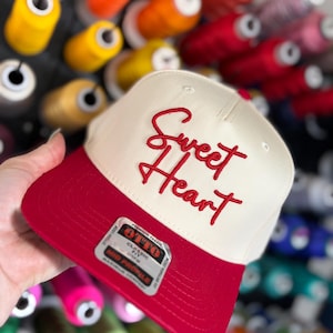 Könnte beinhalten: Cremefarbene Baseballkappe mit rotem Schirm und gesticktem Text "Sweet Heart". Die Kappe hat eine klassische Passform und ein mittleres Profil. Im Hintergrund sind bunte Garnrollen zu sehen.