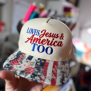 May include: A cream-colored baseball cap with the embroidered text "LOVES Jesus & America TOO" in red and blue. The brim features a floral and American flag design.