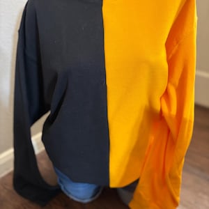 May include: A two-toned sweatshirt, half black and half golden yellow, is displayed on a mannequin. The long-sleeved top is a crew neck style, with a simple, clean design. The color-blocked garment is a casual piece of apparel.