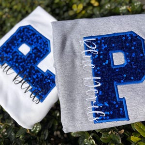 Pode incluir: Duas camisolas, uma branca e outra cinzenta, cada uma com uma grande letra "P" azul com lantejoulas. A camisola branca tem o texto "Blue Devils" em itálico. A camisola cinzenta tem o texto "Blue Devils" em itálico.