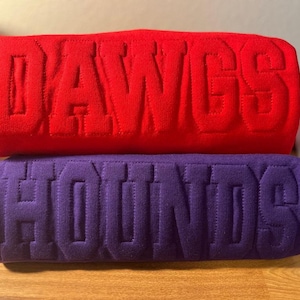 May include: A red sweatshirt with the word DAWGS embossed in large block letters stacked on top of a purple sweatshirt with the word HOUNDS embossed in large block letters. Both garments feature a textured, raised fabric design.