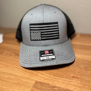 May include: A gray and black trucker hat with a black and white American flag embroidered on the front. The hat has a black mesh back and a label that reads "EST. 1970 RICHARDSON. ORIGINAL 112 TRUCKER ADJUSTABLE".