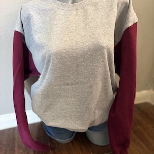 May include: A gray sweatshirt with maroon sleeves. The crew neck sweatshirt is displayed on a mannequin. The sleeves are a solid maroon color, while the body of the sweatshirt is a heather gray.