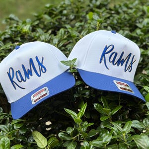 May include: Two white baseball caps with blue brims and the word "Rams" embroidered in blue script. The caps are displayed against a backdrop of green foliage, showcasing the hats' design and color contrast.