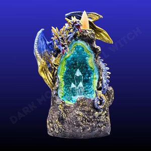 May include: A dragon-shaped incense burner in blue, gold, and green. The design includes a geode-like centre with crystals, framed by the dragon's wings and body. The base has a textured appearance, and the overall design is highly detailed.