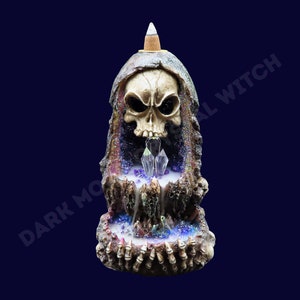 May include: A ceramic incense burner shaped like a skull with a hooded robe. The skull has a detailed face with glowing eyes and a crystal-like structure in the mouth. The base of the burner is decorated with bones and crystals.