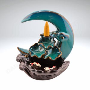 May include: A teal blue ceramic incense burner shaped like a crescent moon with a three-tiered lotus flower design. The burner has a small cone incense holder and three small fish figurines in the bottom tier.