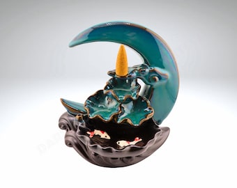 May include: A teal blue ceramic incense burner shaped like a crescent moon with a three-tiered lotus flower design. The burner has a small cone incense holder and three small fish figurines in the bottom tier.