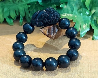 May include: Black beaded bracelet with a carved jade charm. The charm is in the shape of a mythical creature with intricate details. The bracelet is made of round, black beads.