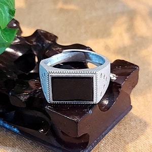 Könnte beinhalten: Ein silberner Ring mit einem schwarzen rechteckigen Stein, der in einer silbernen Fassung sitzt. Der Ring befindet sich auf einem dunkelbraunen Holzständer.